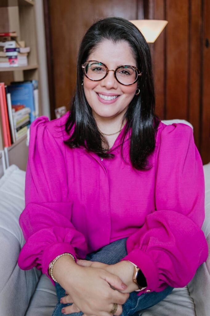 Camila Almeida, psicóloga, vestida com uma camisa rosa e calça jeans, sentada numa poltrona em seu consultório.